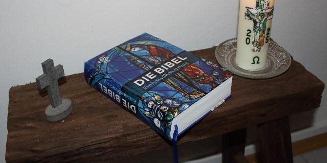 Zu sehen ist ein kleiner Holztisch mit einer brennenden Kerze, einer Bibel und ein kleines Kreuz