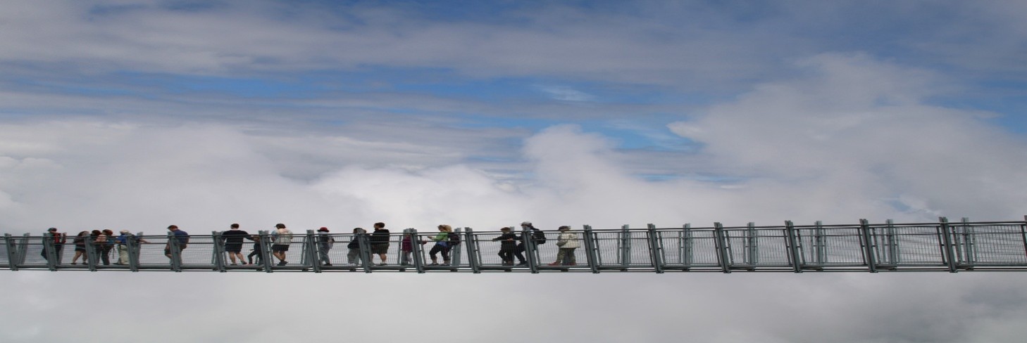 Zu sehen ist eine Hängebrücke mit Menschen vor einer Wolkenkulisse
