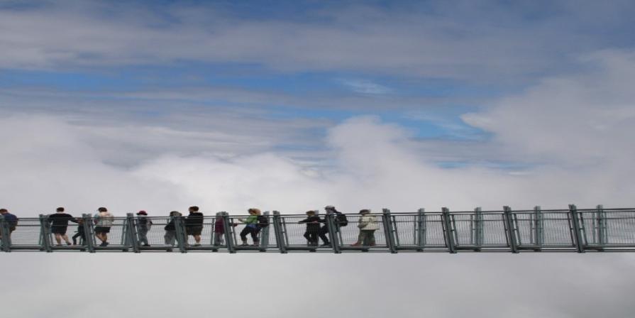 Zu sehen ist eine Hängebrücke mit Menschen vor einer Wolkenkulisse